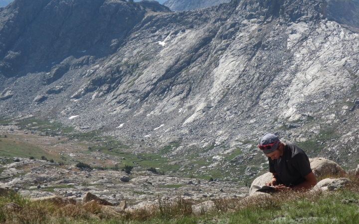 A person sits on a rock and journals. There is a mountain in the background.